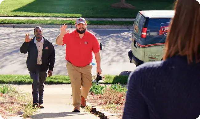 Two men, one in a red shirt and hat and the other in a dark jacket, wave while walking up a sidewalk toward a person standing at the door. A van with “Plumber” branding is parked nearby.